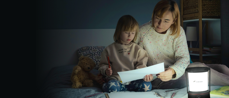 Mother sitting with daughter on bed looking at a piece of paper during a power outage with a lantern helping to light the room